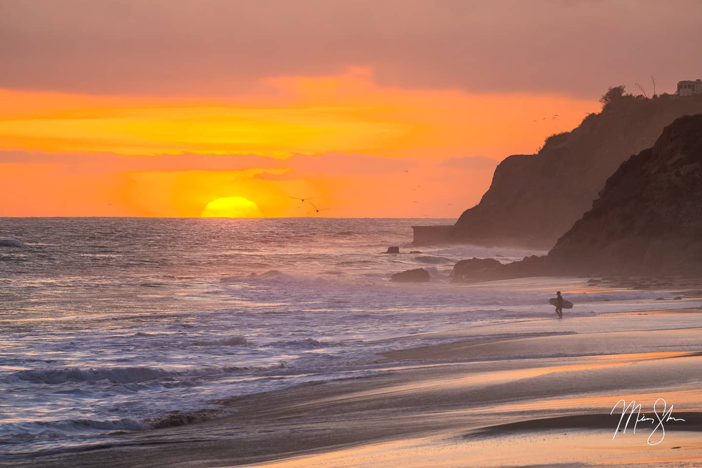 A single surfer contemplates the waves at Leo Carillo State Beach in Malibu as the sun sets in the background. The cliffs and crashing waves add to the drama of the beautiful sunset as a pair of seagulls fly by.