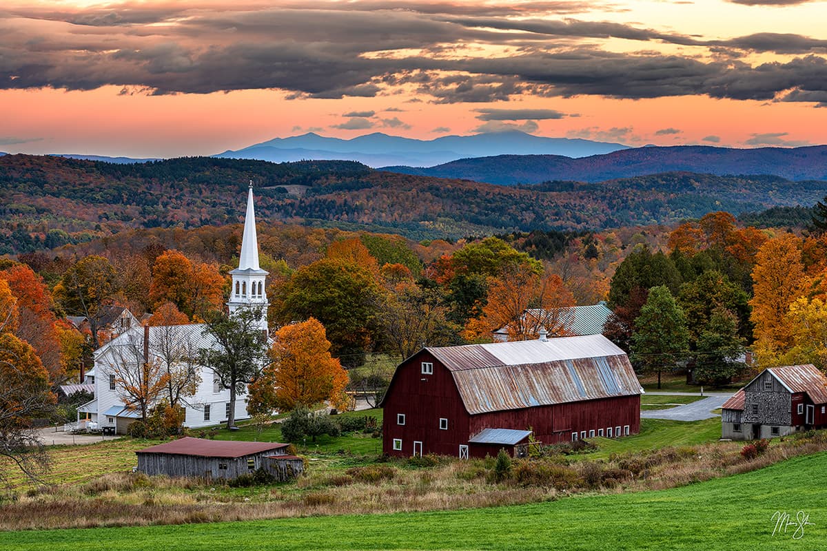 Panorama of Peacham Vermont with historic white church steeple and red barn surrounded by autumn foliage at sunset