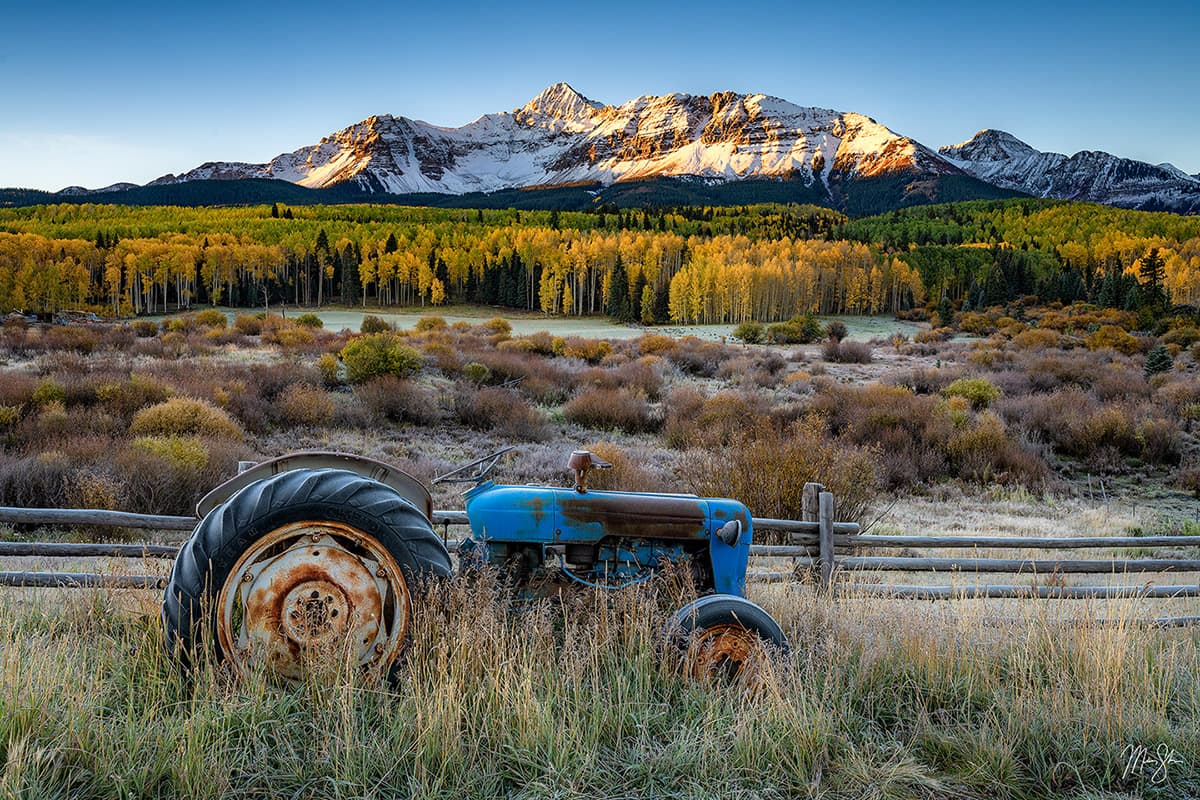 Fine art landscape photography of a vintage tractor at sunrise with the snow-capped Wilson Range in the background near Telluride, Colorado.